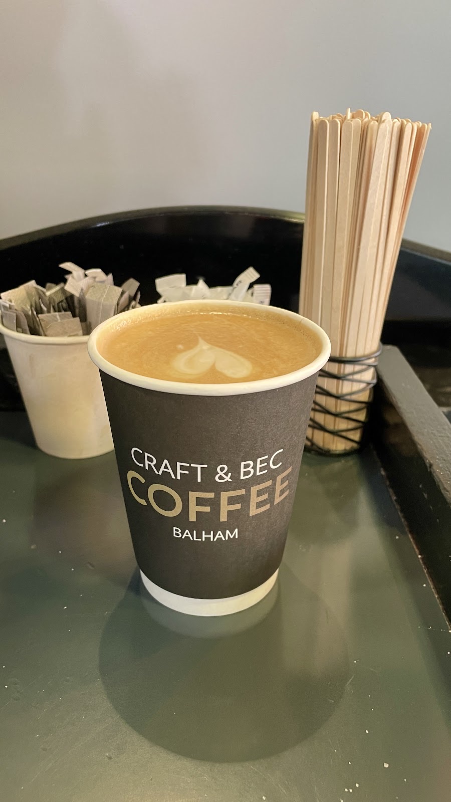 A small coffee with a heart in the foam on a tray beside wooden stirrers and sugar sachets
